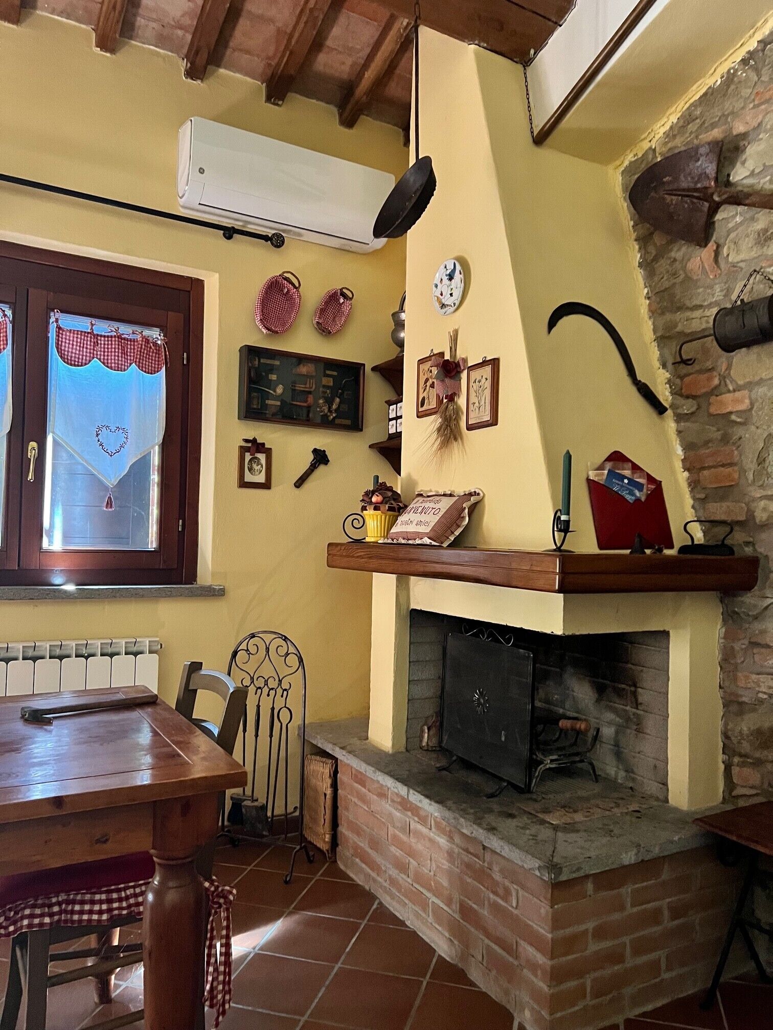 Ein rustikales Interieur mit Kamin aus Backsteinen, einem Esstisch und einem Fenster mit kariertem Vorhang.