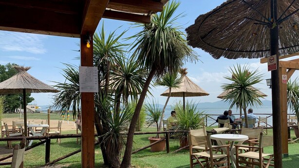 An outdoor seating area with tables and chairs, surrounded by palm trees and a view of the sea. - Holiday Home 'Il Forcone' with Shared Pool, Wi-Fi and Air Conditioning (Panicale)