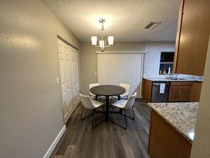 Une cuisine compacte avec un coin repas, comprenant une table ronde avec des chaises, un four encastré et une fenêtre avec des stores.