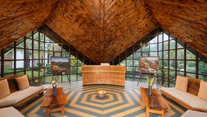 Amplia habitación con techo alto, paredes de madera y ventanas grandes que ofrecen vistas al exterior. Hay dos sofás y una mesa de centro en el centro, con cuadros sobre caballetes apoyados contra la pared.