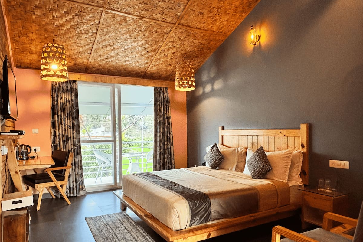 A bedroom with a wooden bed, a desk, and a chair. There is a window with curtains and a view of greenery outside.