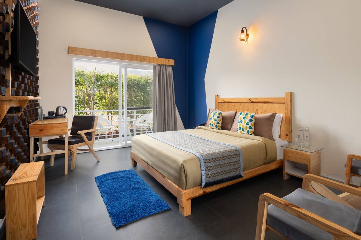 A bedroom with a bed, a desk, a chair, a blue rug, and a balcony with a view of greenery.