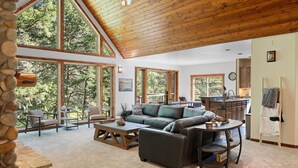 A spacious living room with a high ceiling, large windows, a fireplace, and a kitchen area. - * * *Waters Edge Lodge* **************** (Bonner-West Riverside)