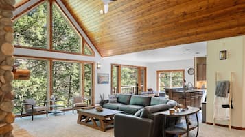 A spacious living room with a high ceiling, large windows, a fireplace, and a kitchen area.