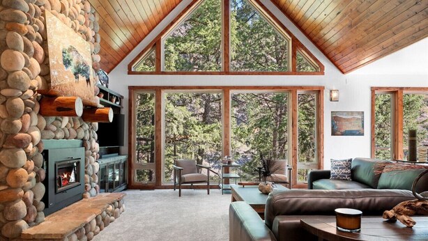 A cozy living room with a stone fireplace, large windows, and a high ceiling. - * * *Waters Edge Lodge* **************** (Bonner-West Riverside)