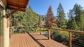 A wooden deck with a railing overlooking a forested area with tall trees and a clear sky. - * * *Waters Edge Lodge* **************** (Bonner-West Riverside)