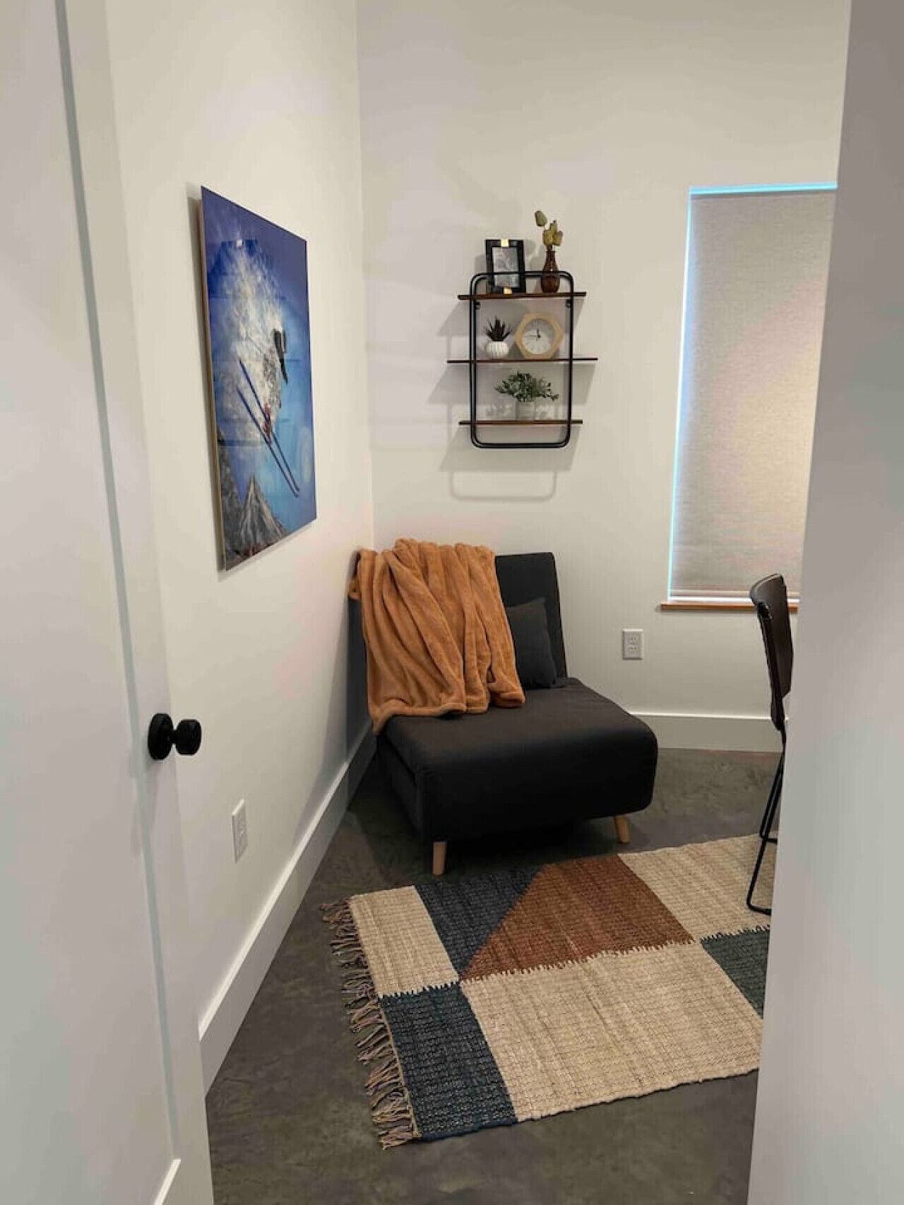 A room with a black sofa, a patterned rug, a wall-mounted shelf with decorative items, and a framed picture.