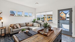 Un salon moderne avec un canapé d’angle, une table basse en bois et de grandes fenêtres donnant sur les bâtiments voisins.