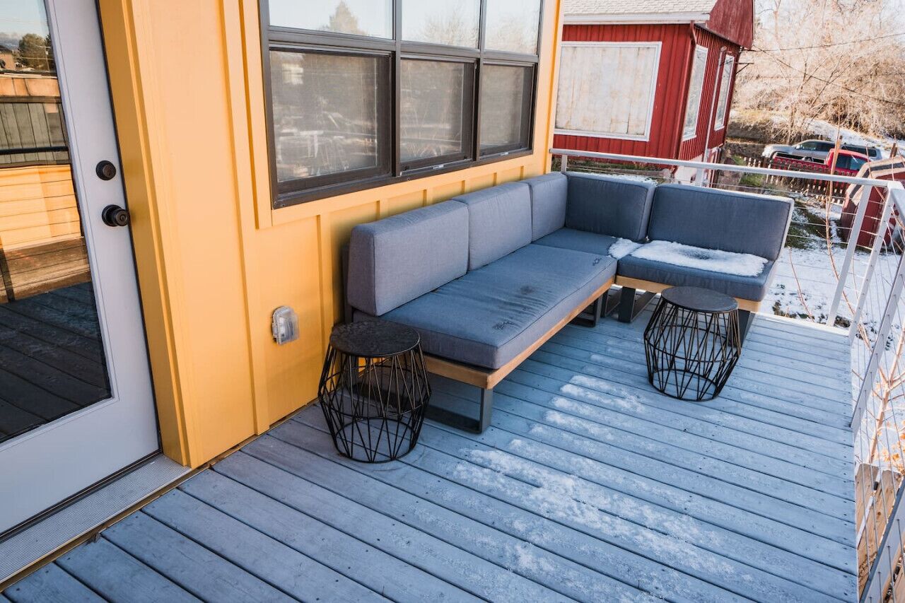 A covered outdoor seating area with a bench and two stools, a door, and a red building in the background.