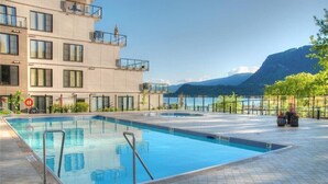 A swimming pool with a diving area, surrounded by a building with balconies and a view of a lake and mountains.