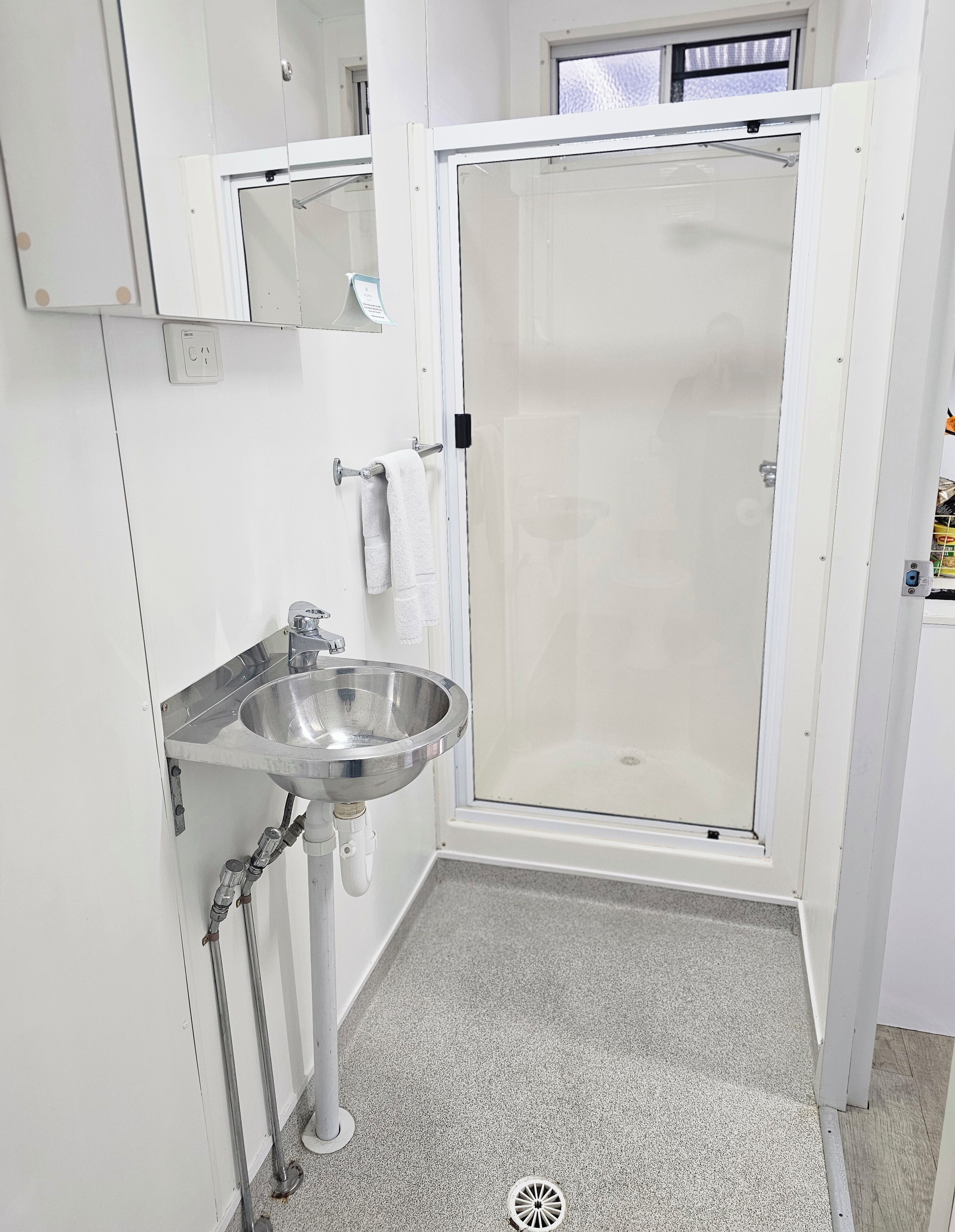 Un baño con un lavamanos de acero inoxidable, una cabina de ducha de vidrio y piso cubierto de baldosas blancas.