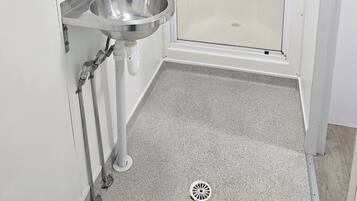 Un baño con un lavamanos de acero inoxidable, una cabina de ducha de vidrio y piso cubierto de baldosas blancas.
