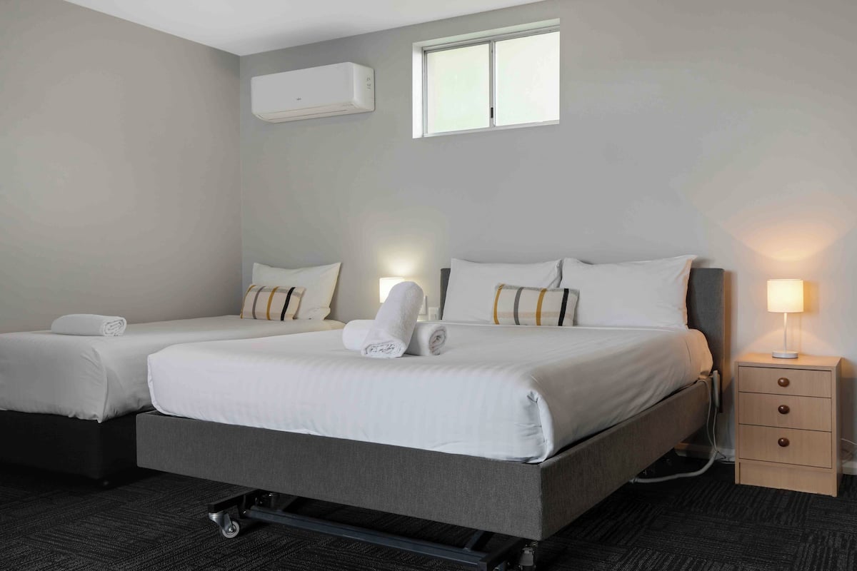 A hotel room with two beds, a small bedside table with a lamp, an air conditioning unit, and a window.