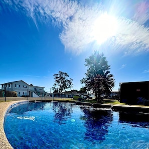 A swimming pool with clear blue water, surrounded by a fence, and a building in the background. - Hive Hotel  (Moruya)