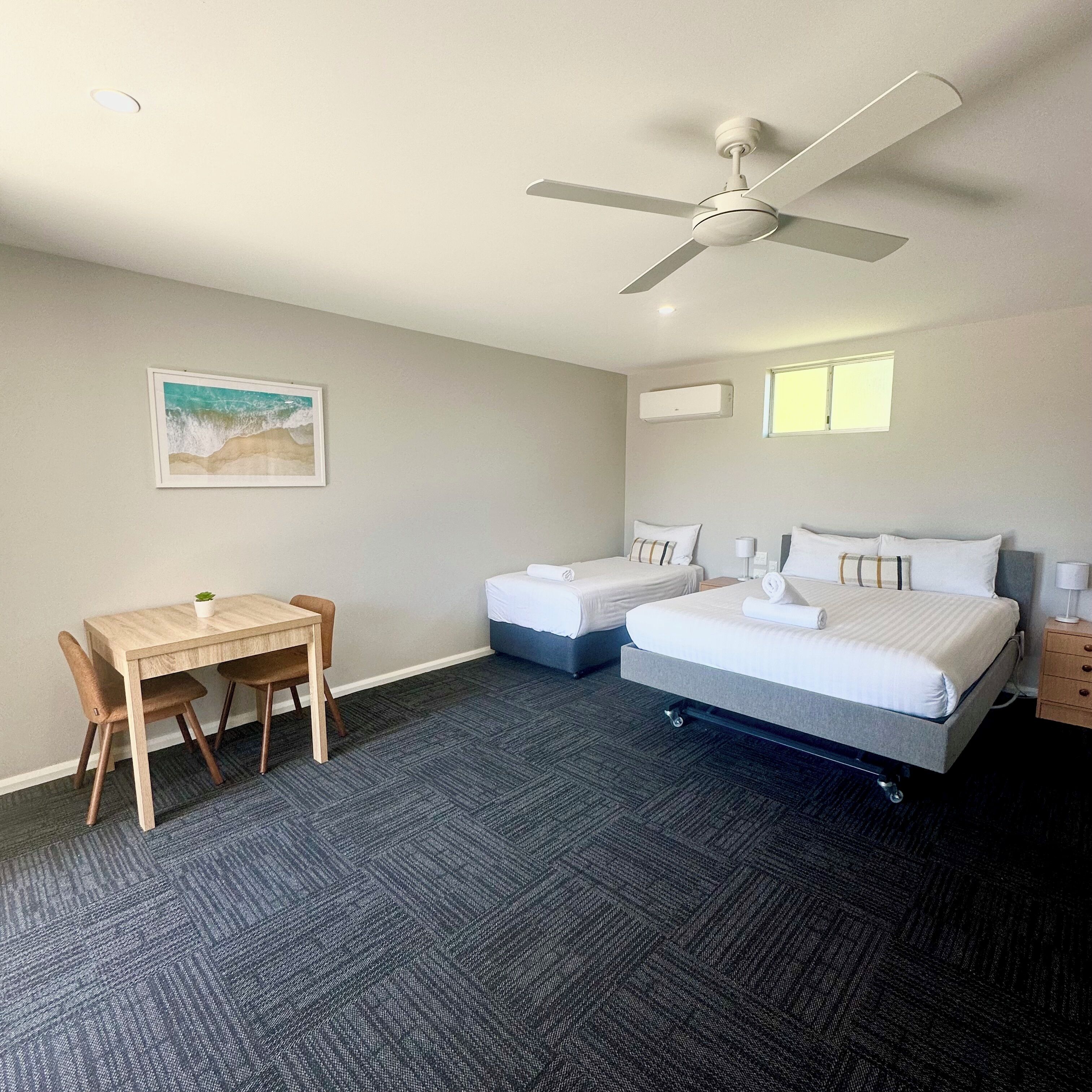 Een hotelkamer met twee bedden, een bureau, een stoel, een plafondventilator en een afbeelding met een strandthema aan de muur.
