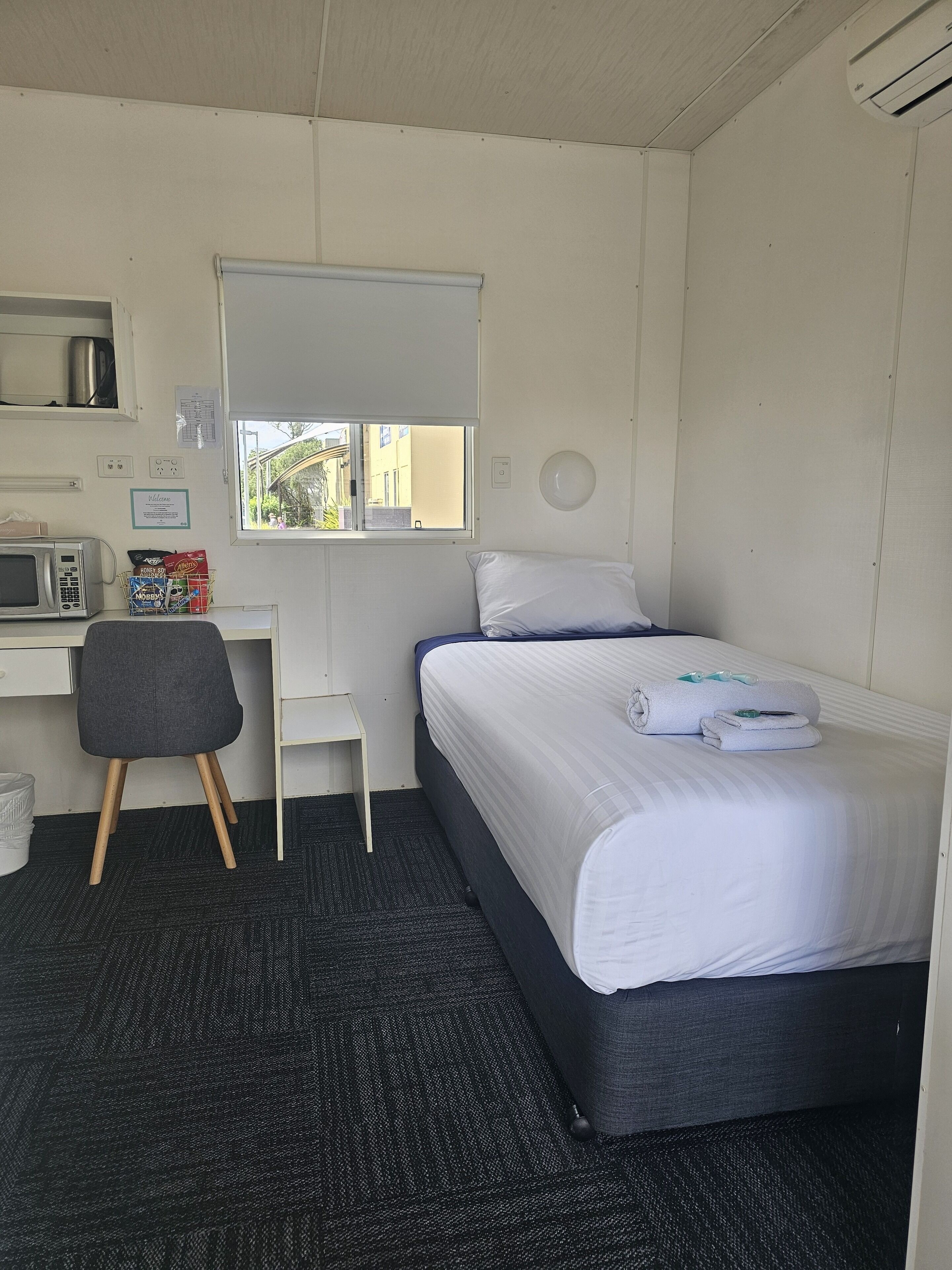 Habitación de hotel compacta con una cama individual, un escritorio pequeño con una silla, un microondas y una ventana con vistas a edificios.