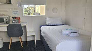 Habitación de hotel compacta con una cama individual, un escritorio pequeño con una silla, un microondas y una ventana con vistas a edificios.