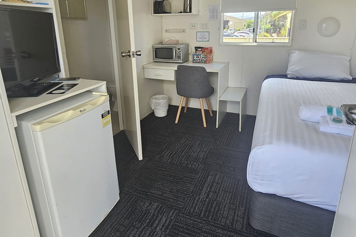 A compact hotel room with a bed, a desk with a computer and a microwave, a small fridge, and a window with a view of the outside.