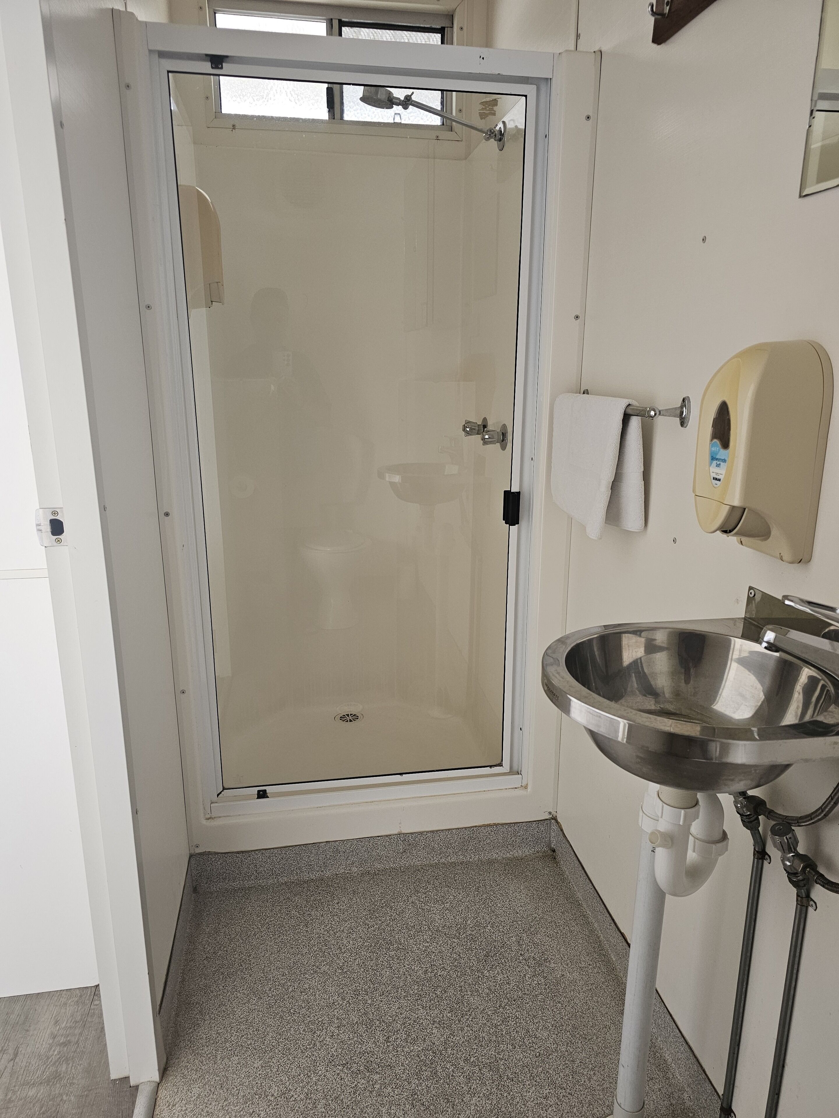 Un baño con cabina de ducha de vidrio, lavamanos de acero inoxidable y dispensador de toallas de papel.