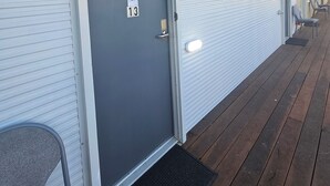 Una hilera de habitaciones de motel con puertas cerradas, letreros con números y un área exterior cubierta con sillas.
