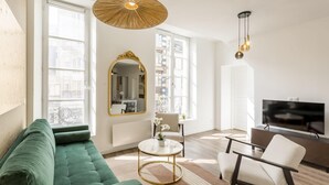 A modern living room with a green sofa, a round coffee table, a white armchair, a TV, and a large mirror. - Merveil - Family Suite - Marais - Fbg St Antoine (Paris)