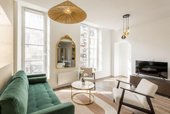 A modern living room with a green sofa, a round coffee table, a white armchair, a TV, and a large mirror.