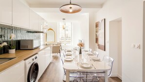 A modern kitchen with a built-in washing machine, a dining area with a white table set for six, and a large window providing natural light. - Merveil - Family Suite - Marais - Fbg St Antoine (Paris)