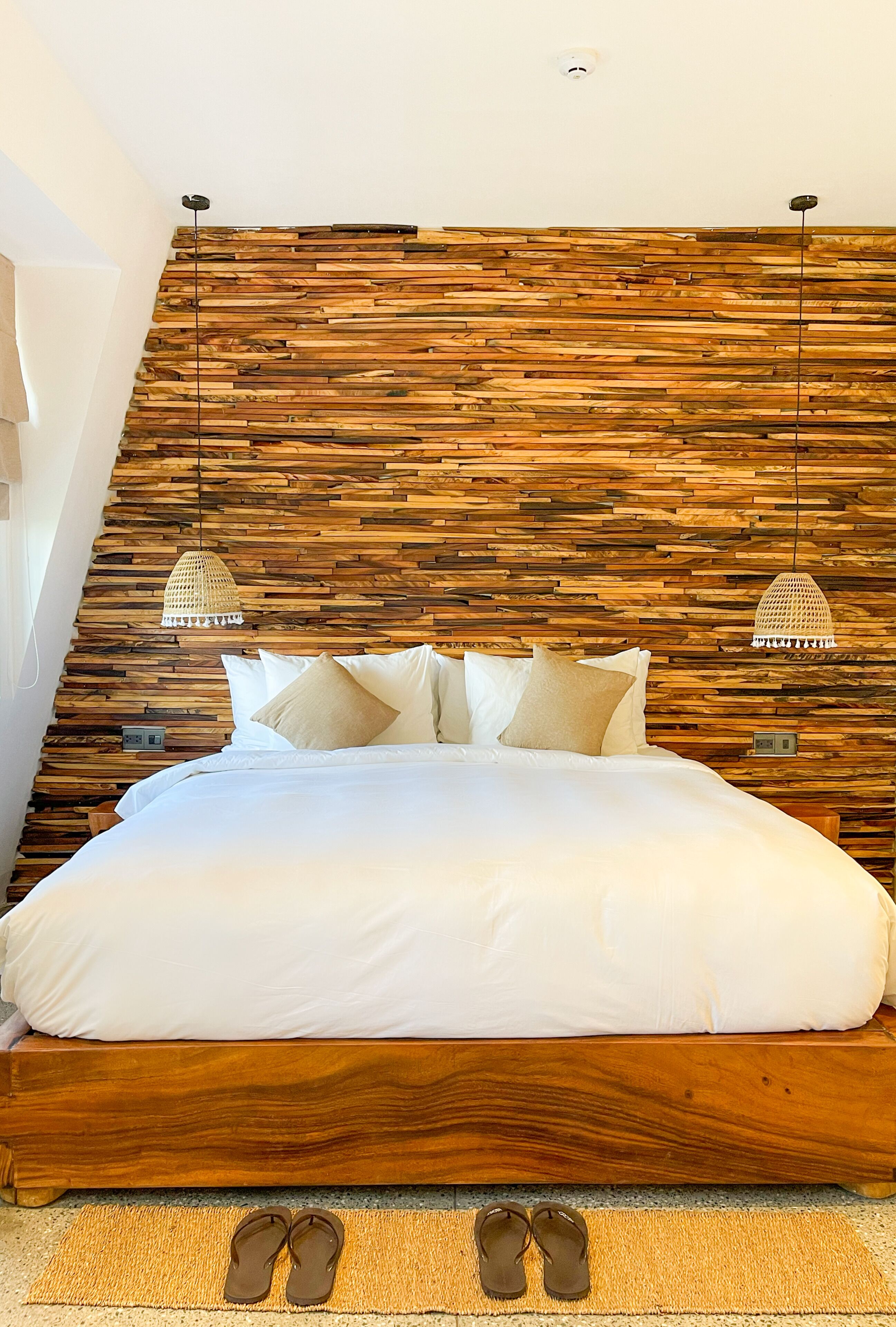 A bedroom with a wooden headboard, a bed with white bedding, two hanging lamps, and a pair of slippers on the floor.