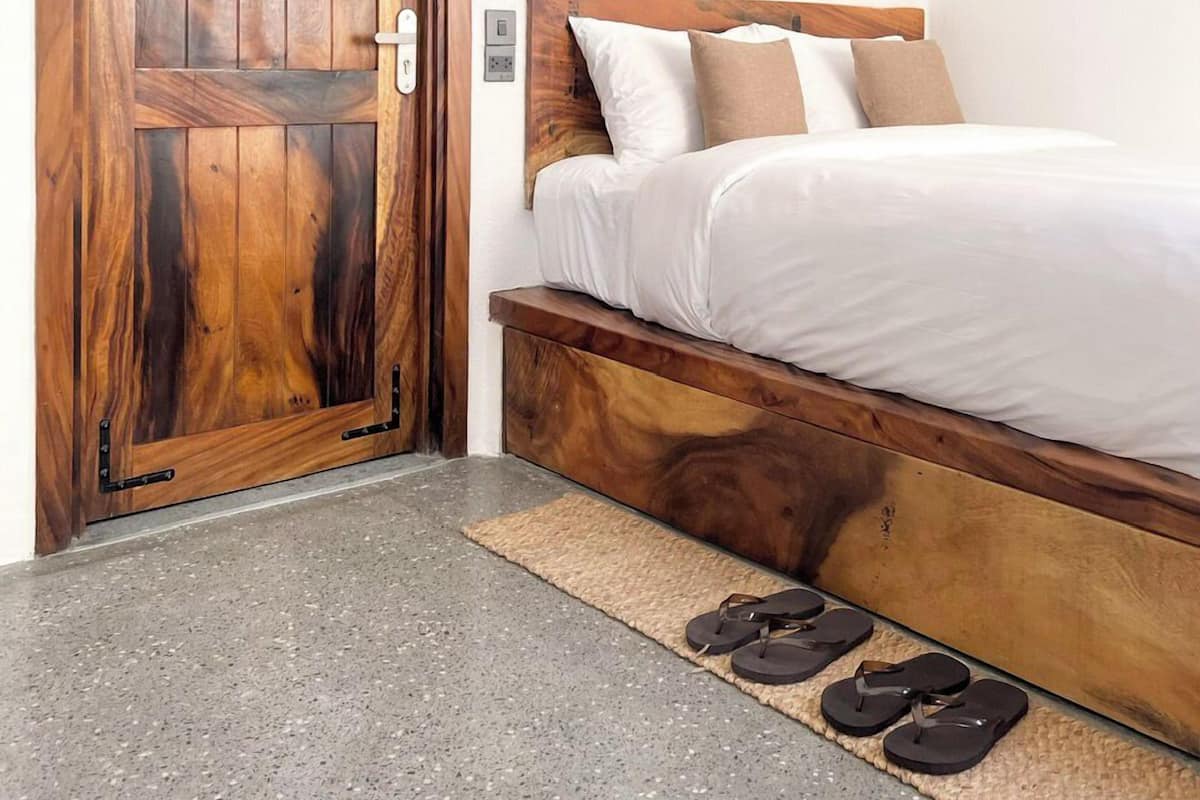 Une chambre avec une porte en bois, un lit recouvert de draps blancs et une paire de tongs posées sur un tapis.