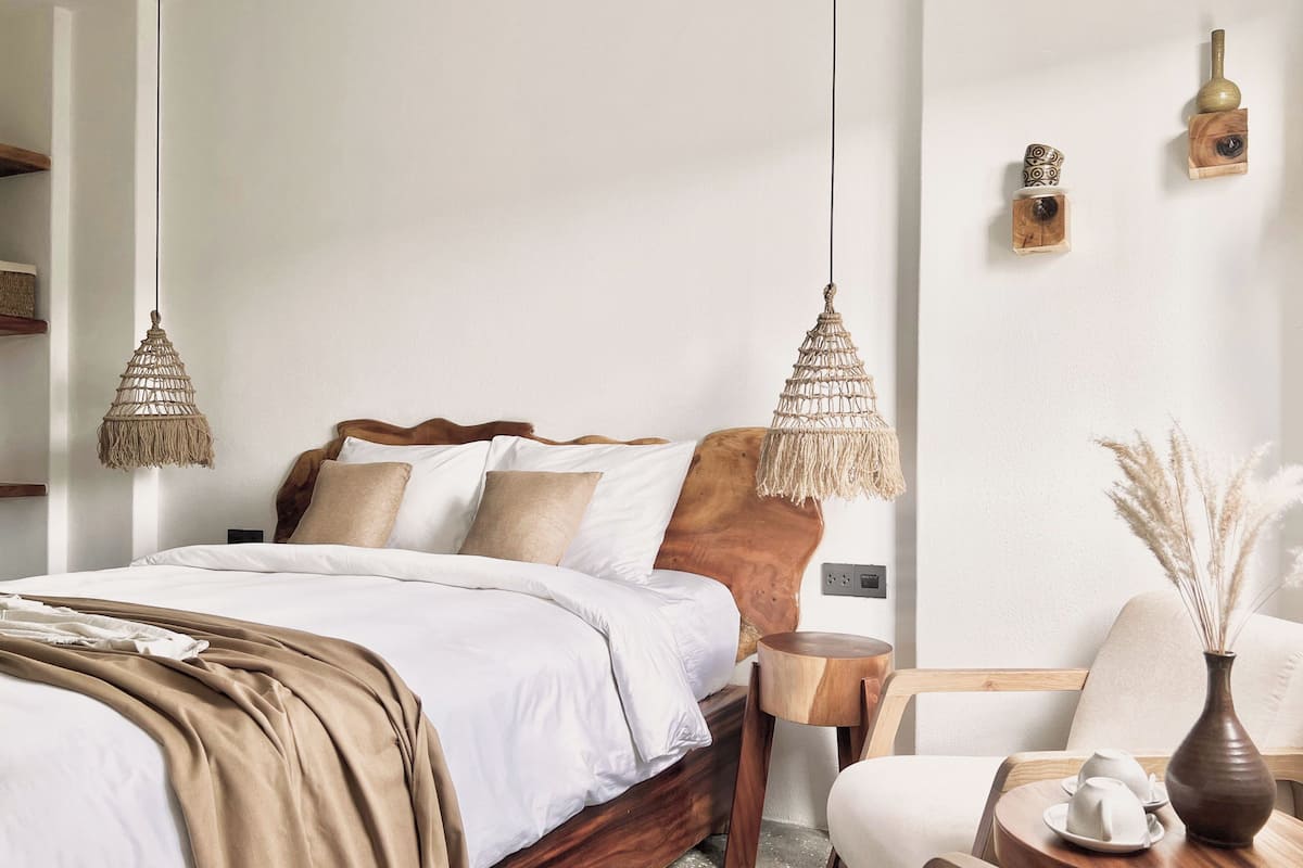 Une chambre à coucher bien rangée, avec un lit en bois, deux lampes en macramé suspendues, une petite table d’appoint en bois, une chaise et un vase contenant des fleurs séchées.