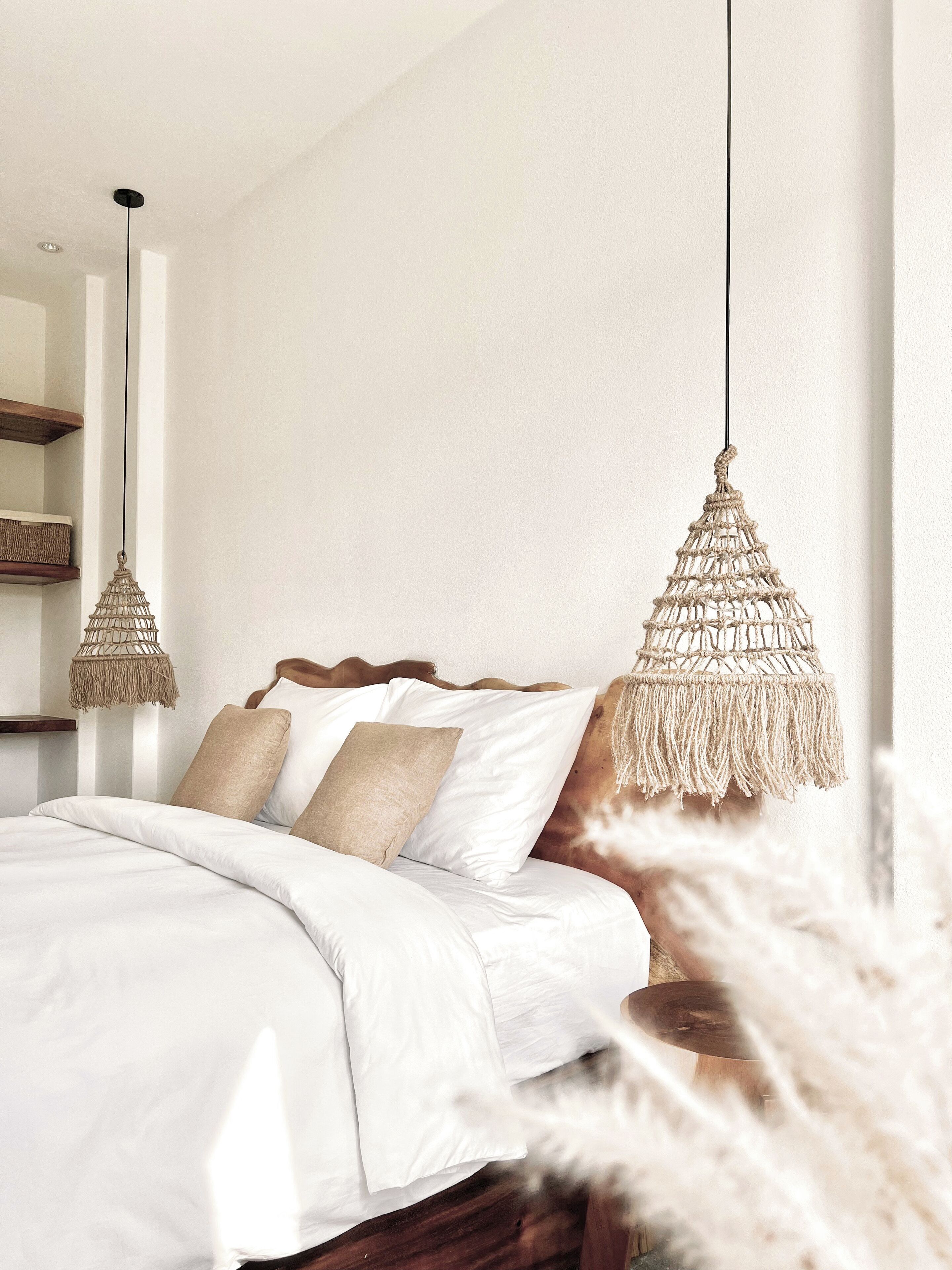 Une chambre à coucher avec un lit, du linge de lit blanc, une tête de lit en bois et deux suspensions décoratives en macramé.