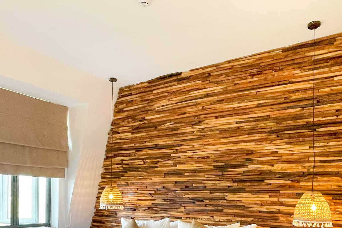 Une chambre à coucher avec une tête de lit en bois, un grand lit recouvert de draps blancs, deux lampes suspendues et une fenêtre avec un store beige.