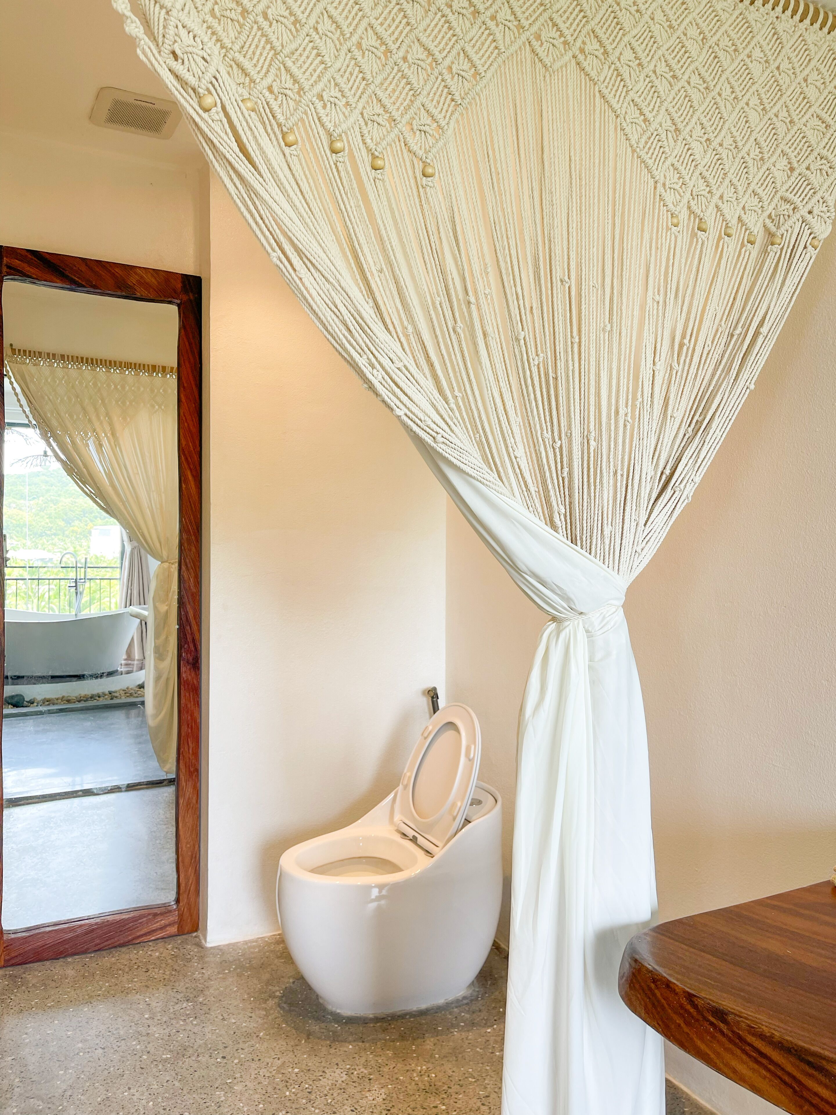 Un baño con cortina de ducha de macramé, inodoro blanco y bañera visible a través de un vano.