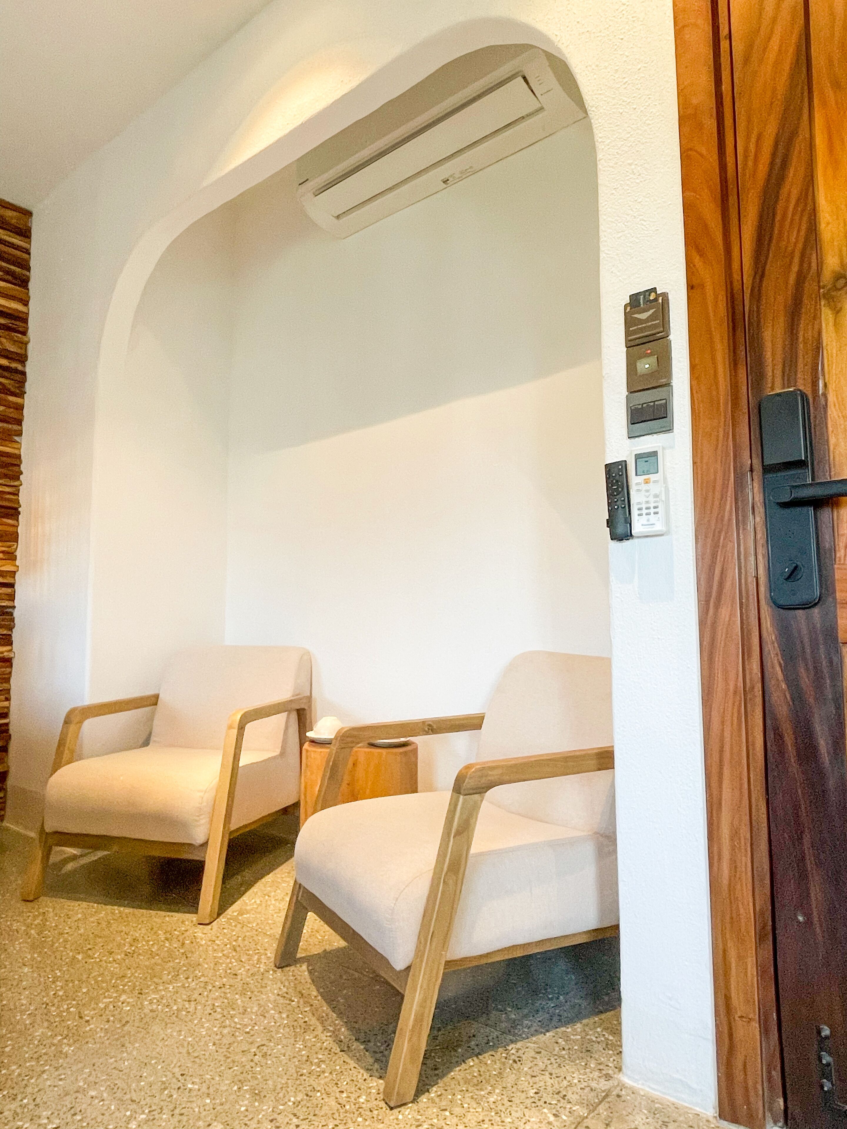 Ein Zimmer mit zwei Holzstühlen, einer Holztür, einer Wand-Klimaanlage und einem kleinen Tisch mit einer Lampe.