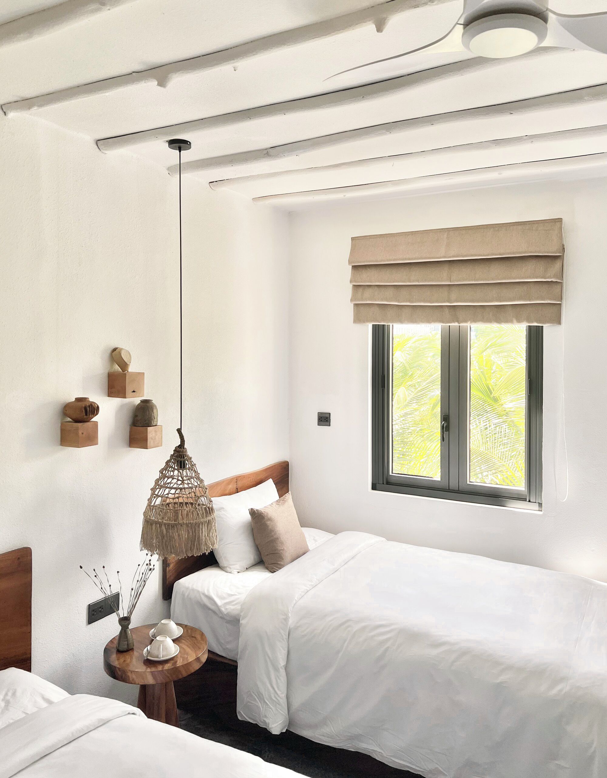 A hotel room with two beds, a window with a view of palm trees, a hanging light fixture, and wall-mounted shelves with decorative items.