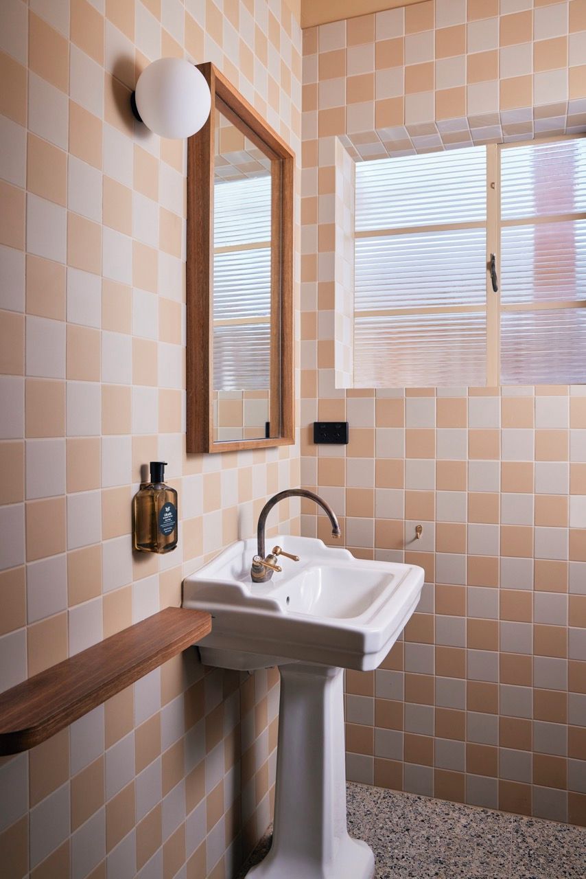 Ein Badezimmer mit rauen Fliesen, einem Standwaschbecken, einem Holzregal, einem Spiegel und einem Fenster mit Jalousien.