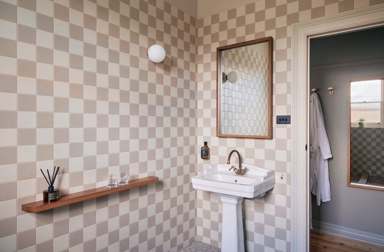 A bathroom with a checkered wall pattern, a pedestal sink, a mirror, and a wooden shelf with toiletries.