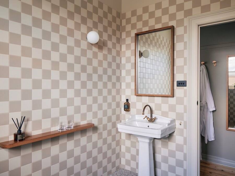 A bathroom with a checkered wall pattern, a pedestal sink, a mirror, and a wooden shelf with toiletries.