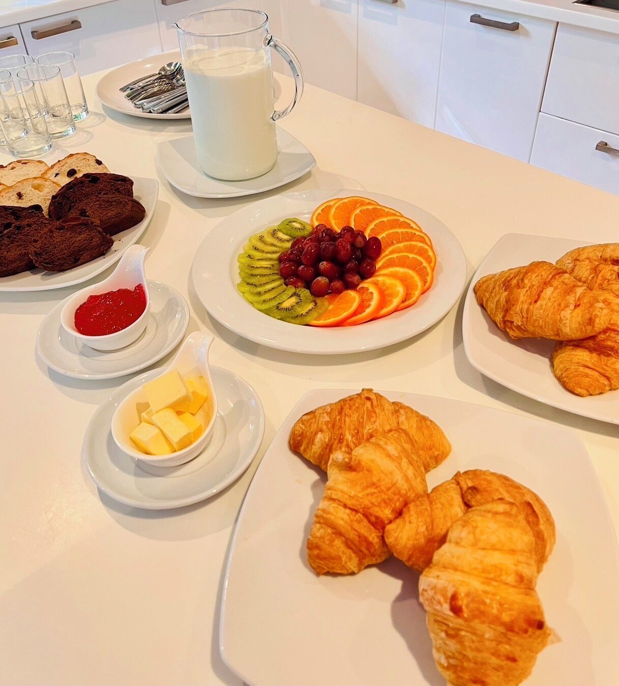 Una mesa de desayuno con croissants, fruta, mantequilla, mermelada y un vaso de leche.
