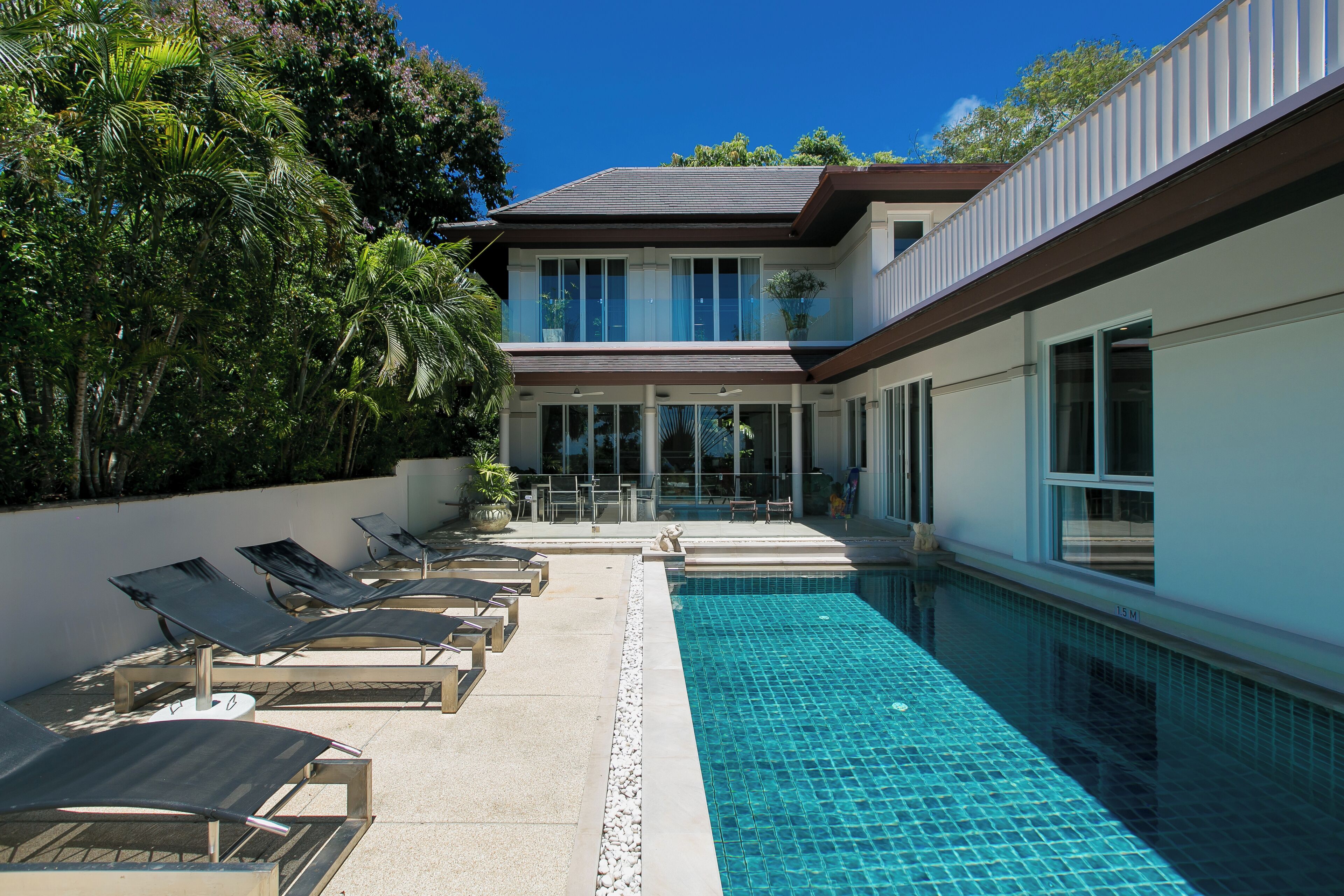 A modern pool area with lounge chairs and a clear blue pool, surrounded by lush greenery and a two-story building with large windows.