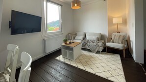 Ein modernes Wohnzimmer mit großem Fenster, Flachbildfernseher, Holzbalke an der Decke, einer Couch, einem Sessel und einem kleinen Tisch mit Lampe.