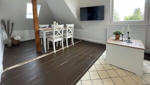 Ein Zimmer mit Holzbalkendecke, einem weißen Esstisch mit Stühlen, einer kleinen weißen Theke und einem großen Fenster.