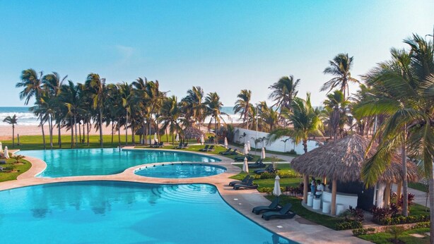 A resort pool with multiple sections, surrounded by palm trees and a beach view.
