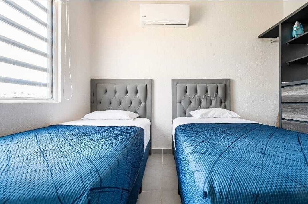Habitación de hotel con dos camas, una ventana con persianas, aire acondicionado y una estantería con un objeto color turquesa.