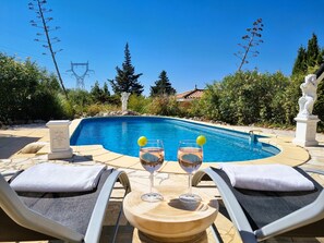 Ein Poolbereich mit zwei Liegestühlen, einem Tisch mit zwei Weingläsern und einer Statue im Hintergrund.