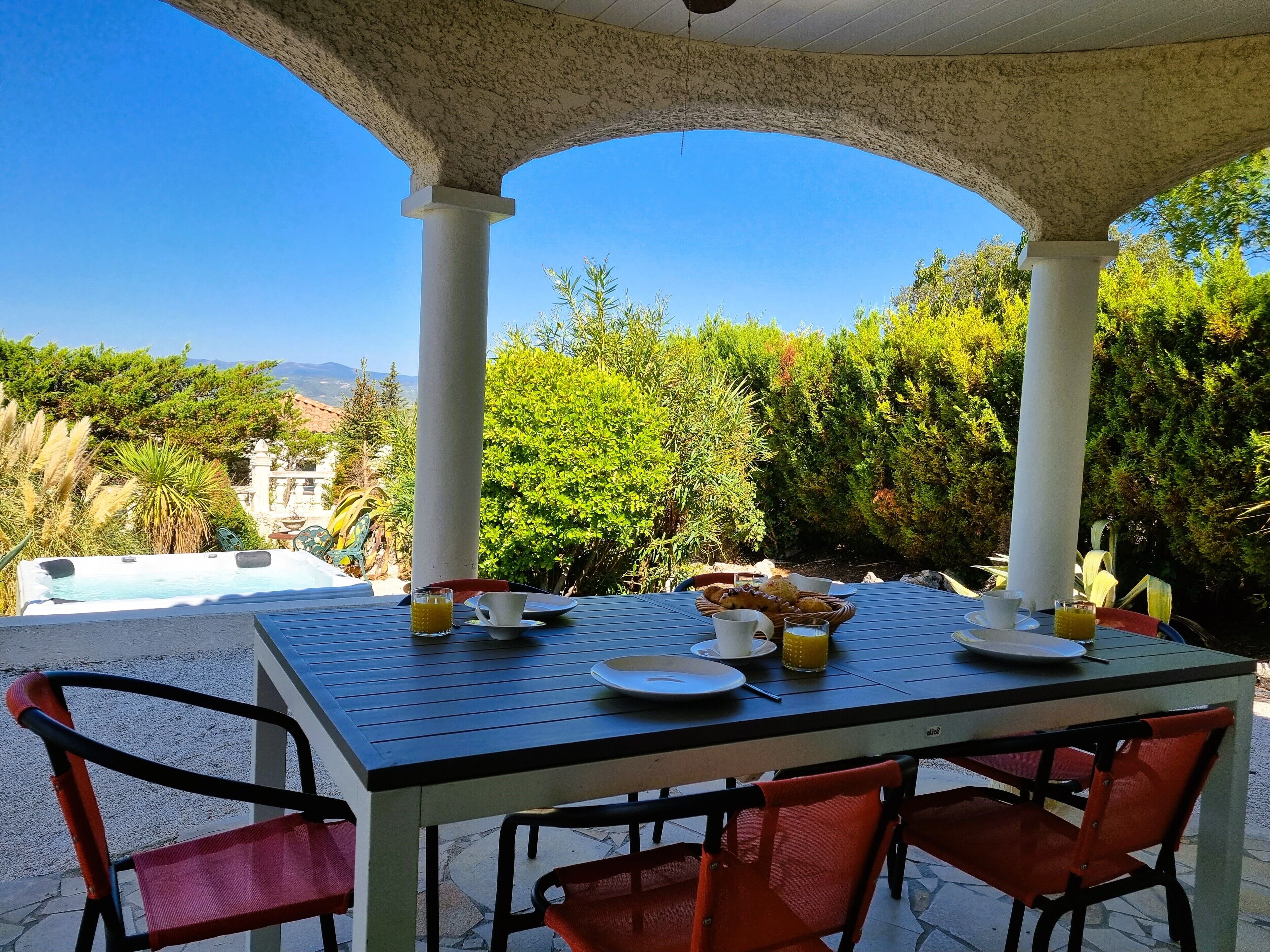 Ein Essbereich im Freien mit einem Tisch für zwei Personen, umgeben von roten Stühlen, unter einer Laube mit Säulen.