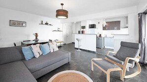Una sala moderna con un sofá gris, una mesa de centro de madera y un sillón gris. Se observa una cocina al fondo con armarios blancos y una zona de bar.