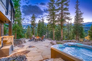 A stone patio with a hot tub, outdoor seating, and a view of mountains and trees. - Shock Hill Overlook Gem | Gondola In/Out | VIEWS! (Breckenridge)