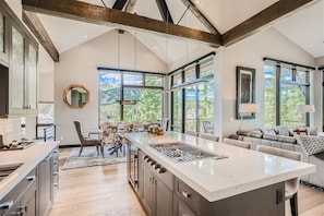 A modern kitchen with a large island, wooden beams, large windows, and a view of the outdoors.