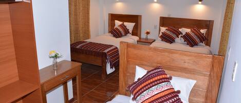 Habitación de hotel con dos camas de madera, cada una con almohadas y cabeceros estampados, una mesita de noche de madera con un florero y un escritorio de madera.